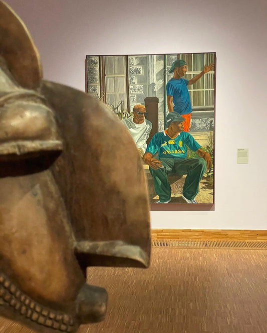 A gallery installation image with a carved wooden mask in the foreground and colourful painting of 3 men by Feni Chulumanco in the background.